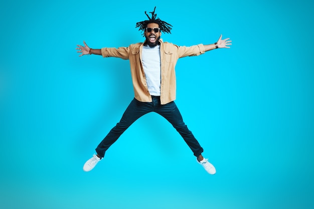 Photo full length of happy young man in casual wear keeping arms and legs outstretched while hovering against blue wall