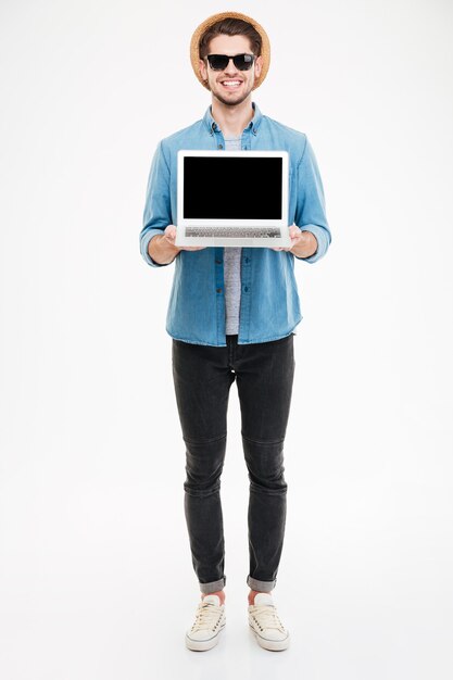 Photo full length of happy young man in sunglasses and hat holding blank screen laptop