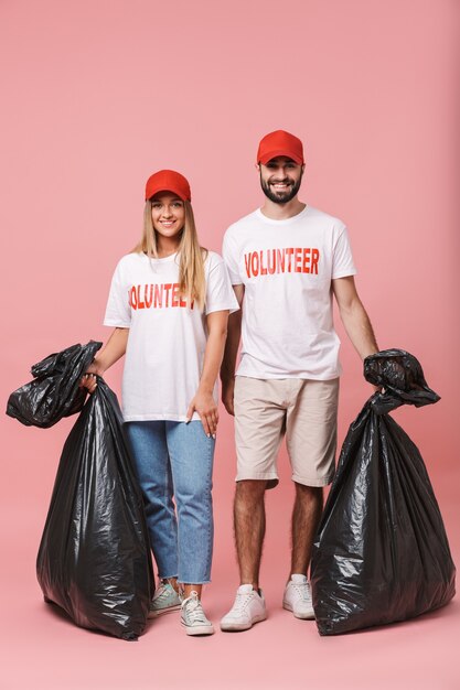 Full length of a happy young volunteers couple standing isolated, holding rubbish bags