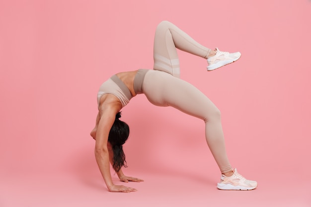 Full length of a healthy fit young woman wearing sportswear doing stretching exercises isolated over pink wall