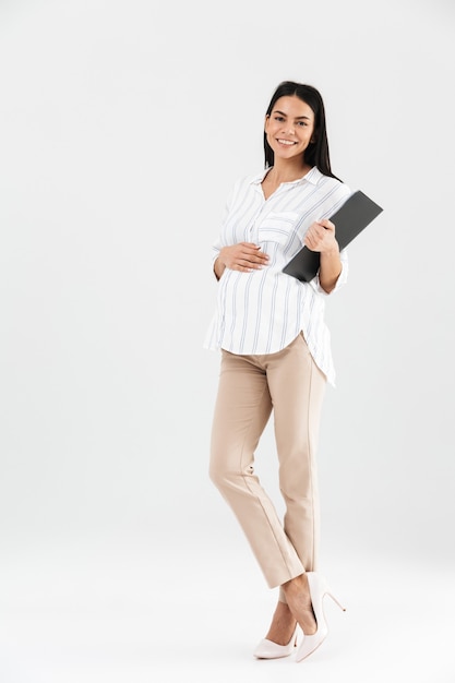 Full length healthy pregnant businesswoman holding clipboard and touching her big tummy while standing isolated over white wall