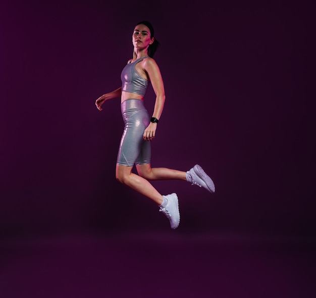 Полная длина здоровой женщины в серебряной спортивной одежде, прыгающей в воздух на фиолетовом фоне