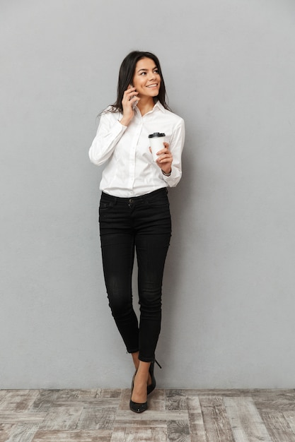 Full length image of beautiful business woman in formal wear having mobile call and looking aside with paper cup of takeaway coffee in hand, isolated over gray background