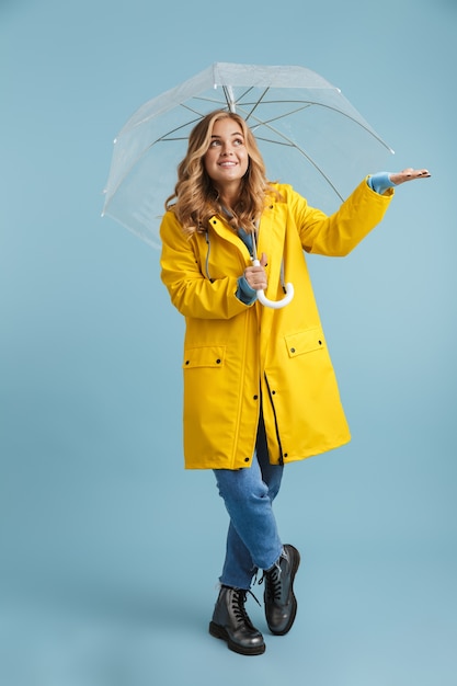 Full length image of blonde woman 20s wearing yellow raincoat standing under transparent umbrella