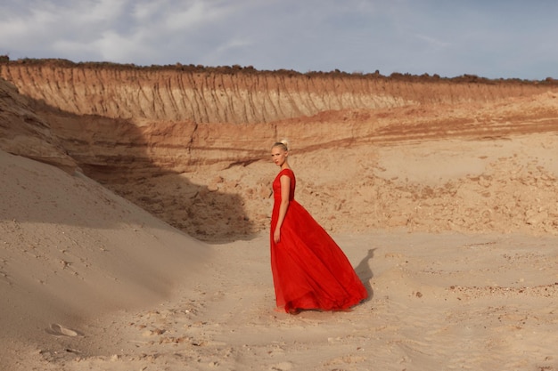 Full length image of a blonde young woman in red waving dress with flying fabric on the background of golden sands.