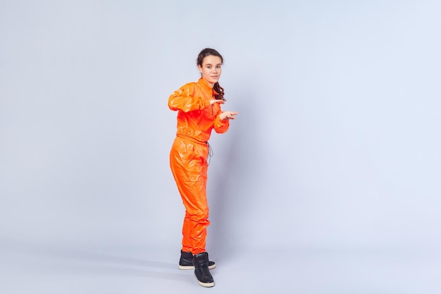 Full length image of cheerful teenage girl with brunette hair wearing bright orange jumpsuit dancing showing hip hop move youth hobby activities indoor studio shot isolated on white background