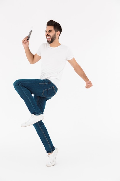 Full length image of excited young man holding cellphone and dancing isolated over white wall