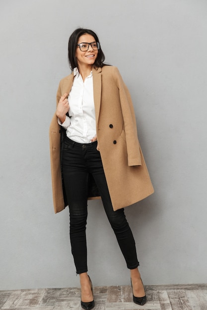 Full length image of fashion woman in glasses wearing heels and beige coat posing at camera with smile, isolated over gray background