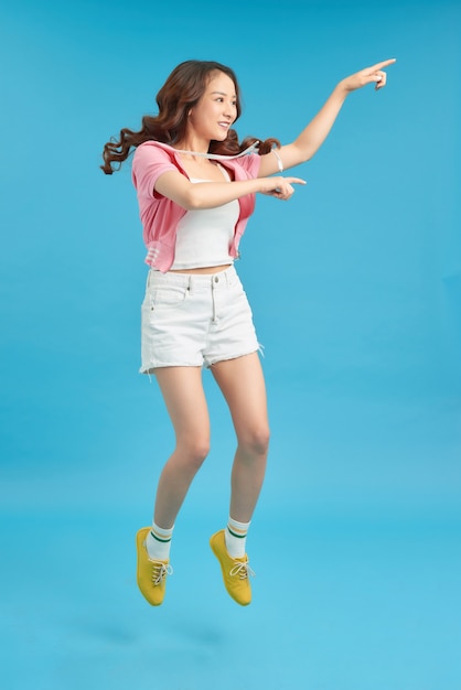 Full length image of Joyful asian woman jumping while pointing and looking away over blue background
