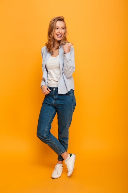 Full length image of pleased woman wearing jeans and sneakers smiling and pointing finger backward on copyspace, isolated over yellow background