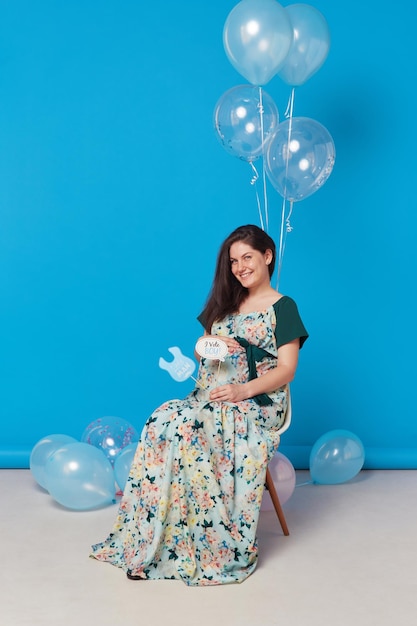 青い背景の上の風船と椅子に座っている妊娠中の陽気なブルネットの若い女性の完全な長さの画像