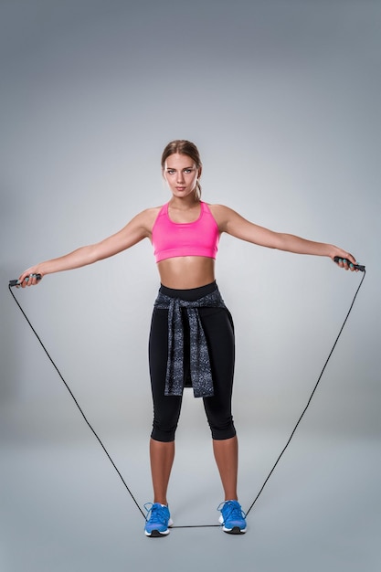 Full length image of a pretty fitness woman doing exercise with skipping rope over gray background. Young woman with beautiful slim healthy body posing in studio.