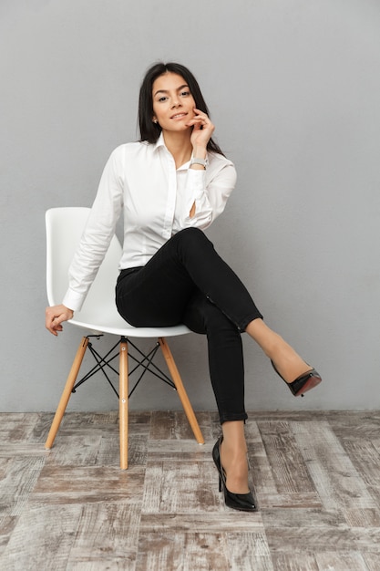Full length image of stylish business woman in white shirt and black trousers sitting on chair in office, isolated over gray background