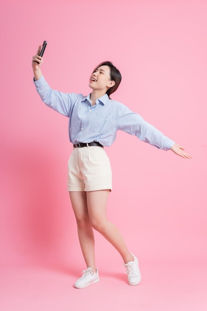Изображение молодой азиатки в полный рост с помощью смартфона на розовом фоне