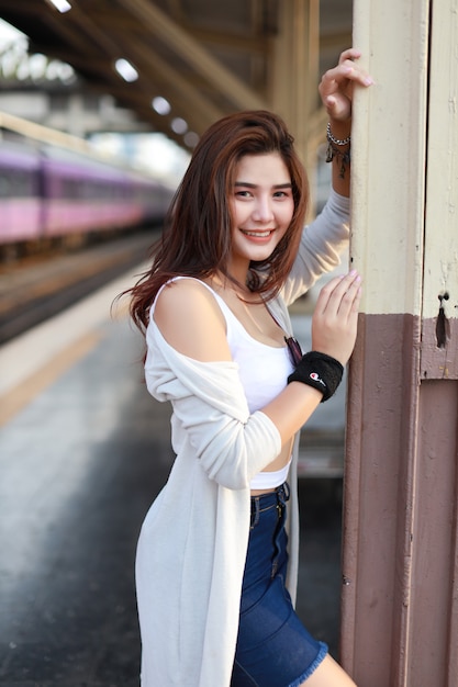 Full length image of young asian woman, long hair in white dress standing and looking at camera while waiting in train station with beauty face