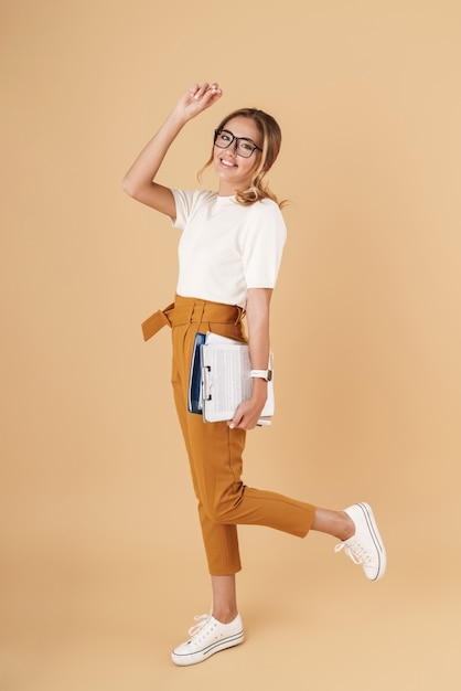 Full length image of young businesswoman wearing eyeglasses holding clipboard and register books isolated