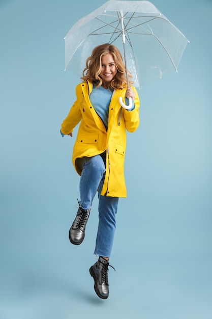 Full length image of young woman 20s wearing yellow raincoat standing under transparent umbrella