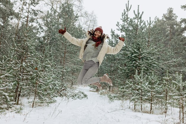 Full length of joyful woman in warm clothes jumping above ground in snowy winter forest
