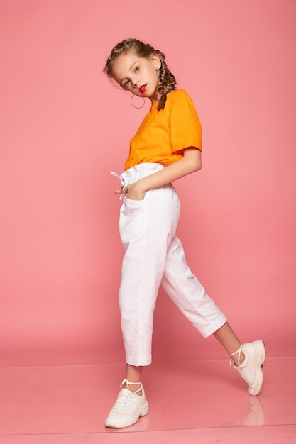ピンクのスタジオの背景にオレンジ色のTシャツと白いズボンと白いスニーカーを着た長身の小さな女の子スタイリッシュな子供幸せな子供時代の感情コピースペース