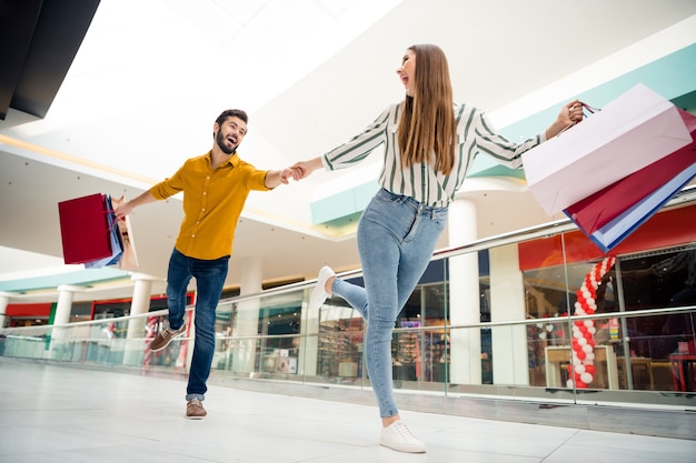 Full length low angle view photo of cheerful pretty lady lead handsome guy hurry next store wanna buy one more shirt dress shoes carry many bags shopping center wear casual outfit indoors