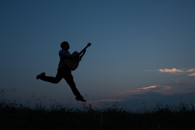Full length of man holding guitar jumping against sky at dusk