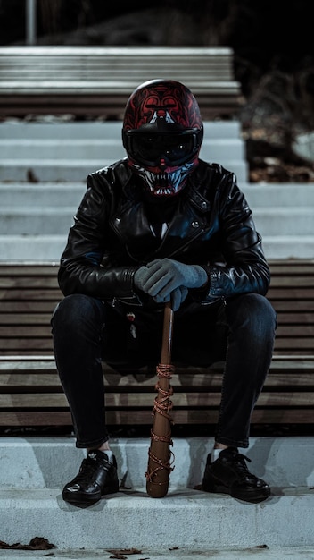 Photo full length of man in a leather jacket and red helmet sitting on bench and holding a baseball bat