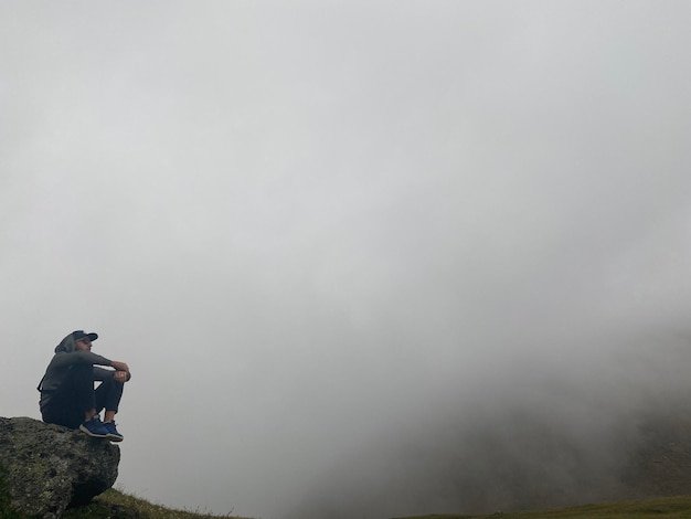 Foto lunghezza intera di un uomo seduto su una roccia contro la nebbia