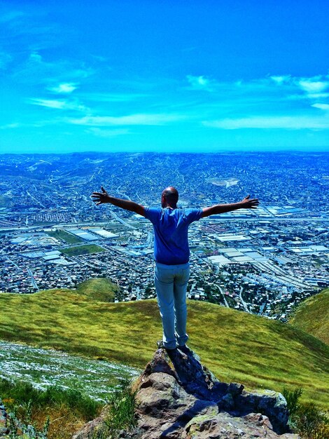 Full length of man with arms outstretched standing on rock over hill against cityscape