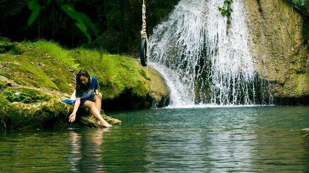 Full length of mature woman touching river water while sitting on rock in forest