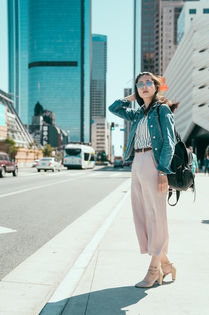 full length modern young woman backpacker travel in big city los angeles. beautiful female tourist standing by road on street waiting taxi commute after visit concert hall music museum in sunglasses