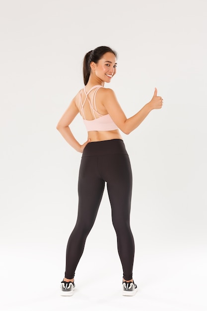 Full length of motivated and satisfied smiling asian female coach, girl athleticin sportswear turn behind to show thumbs-up , ready for productive fitness training, white wall.