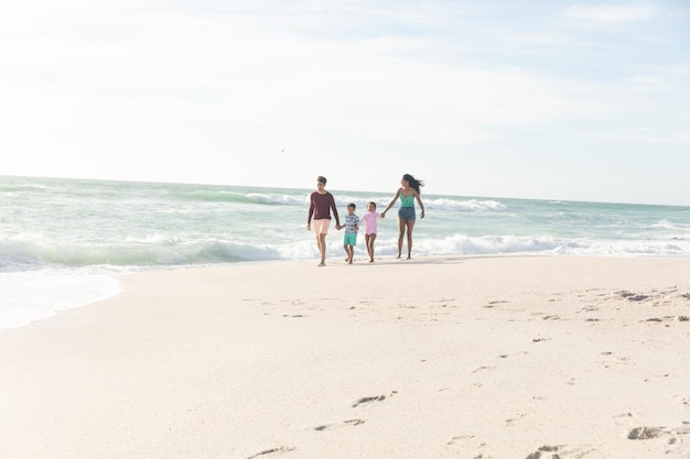 Full length of multiracial family walking on shore holding hands at sunny beach against sky