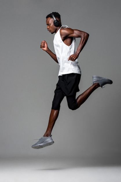 Фото Мускулистый африканский спортсмен в полный рост в спортивной одежде и наушниках слушает музыку и бегает во время тренировки на сером фоне