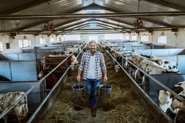 Фото Во всю длину красивого кавказского фермера в клетчатой рубашке и джинсах держит ведра в руках с кормом для животных. стабильный интерьер.