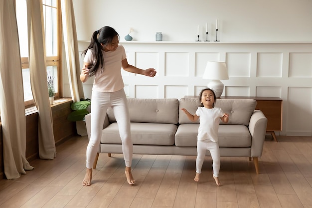 Full length overjoyed different generations asian family in\
home wear enjoying dancing in barefoot together in modern living\
room laughing young vietnamese mother showing moves to little\
daughter