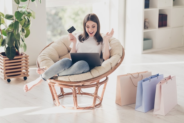 Foto la foto a tutta lunghezza della ragazza stupita si siede sulla sedia guarda il monitor del laptop tiene la carta di credito in casa al chiuso shopping concept di vendita