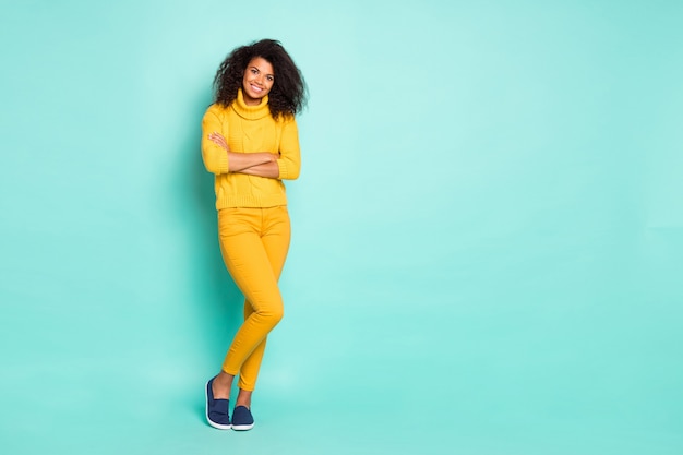 Full length photo of amazing dark skin lady holding arms crossed cool street look wear yellow knitted pullover trousers isolated blue teal color wall 