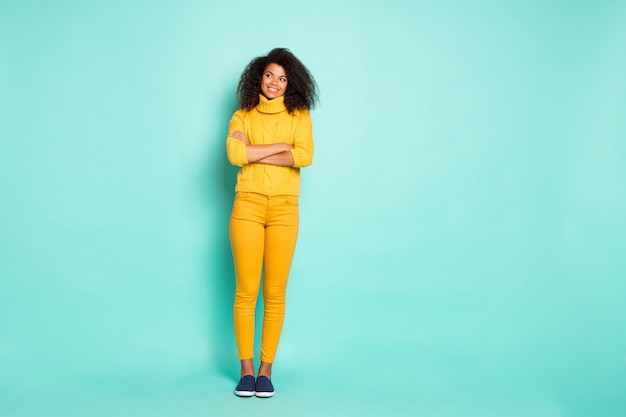 Full length photo of amazing dark skin lady holding arms crossed looking wondered empty space wear yellow knitted pullover trousers isolated blue teal color wall 