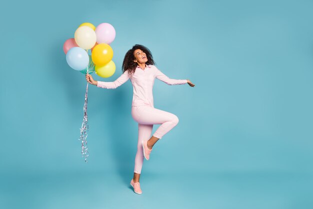 Full length photo of amazing wavy dark skin lady holding many air balloons excited positive party mood wear pink shirt trousers isolated blue color background