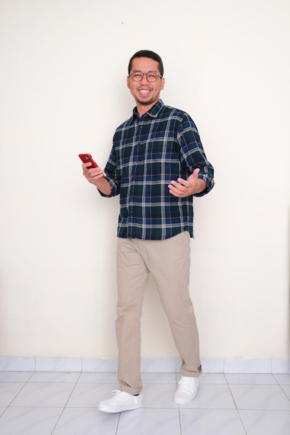 Full length photo of Asian man walking and holding mobile phone with happy expression