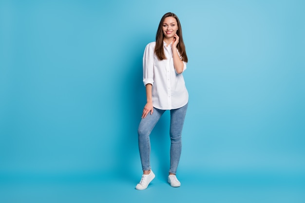 Полная длина фото привлекательной леди хорошее настроение длинная прическа носить белую рубашку джинсы кроссовки изолированный синий цвет фона