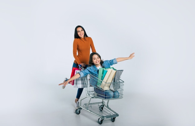 Full-length photo of attractive woman in casual clothes, who is pushing a shopping cart with her daughter, who is pointing forward, and three shopping bags in it.