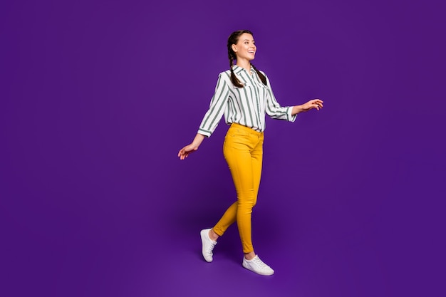 Full length photo of beautiful pretty lady toothy beaming smiling going down street good sunny day weather wear striped shirt yellow trousers isolated purple color background