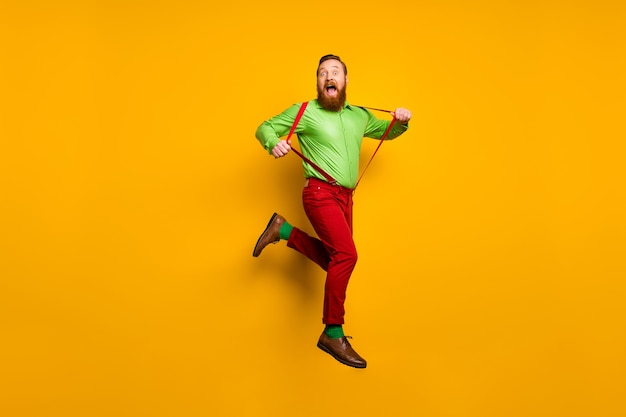 Photo full length photo of careless childish crazy funky man jump pull his modern suspenders scream feel rejoice wear stylish outfit shoes isolated over yellow color