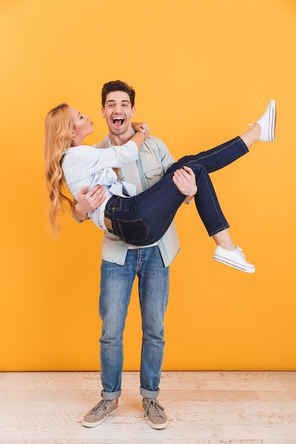 Full length photo of cheerful man smiling and holding woman in his arms who kissing his cheek, isolated over yellow wall