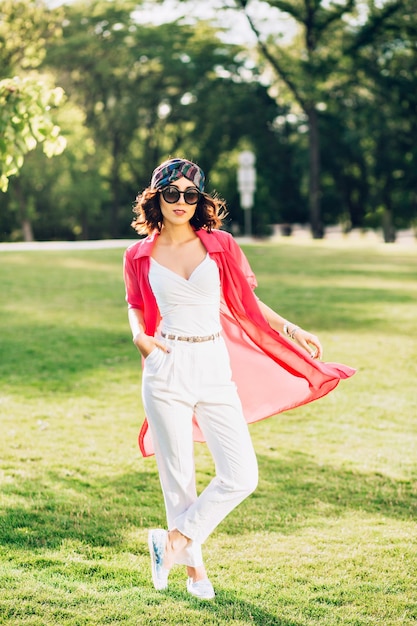 Full-length photo of cute brunette girl in bandana standing in summer park. She wears white clothes, sunglasses, her long pink shirt is flying in sunlight.