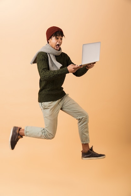 Foto integrale del ragazzo afroamericano eccitato che indossa cappello e sciarpa che cammina con il computer portatile, isolato sopra la parete beige