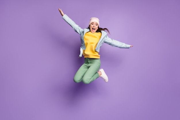 Full length photo of funky attractive lady stylish clothes jump high rejoicing flying arms like wings bird crazy wear casual hat jacket green pants shoes isolated purple color background