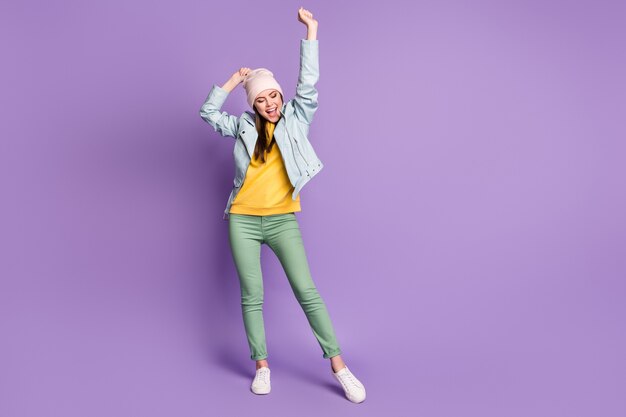 Full length photo of funny attractive lady  good mood raise arms up excited cheerful wear casual hat modern blue jacket green pants sneakers isolated purple color background