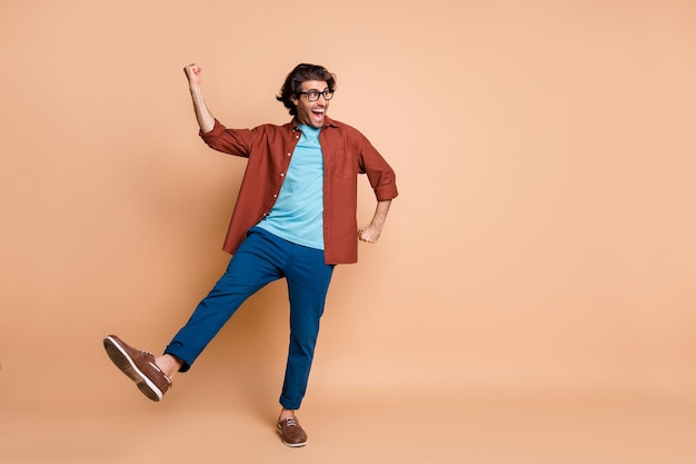 Full length photo of guy dance open mouth raise fist leg wear specs brown t-shirt trousers footwear isolated beige color background